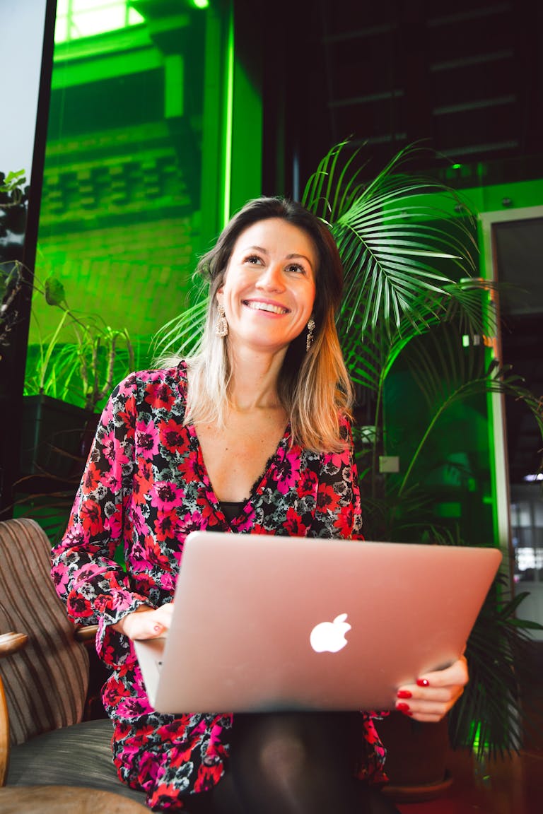 Smiling woman using a laptop in a modern indoor setting with lush greenery.