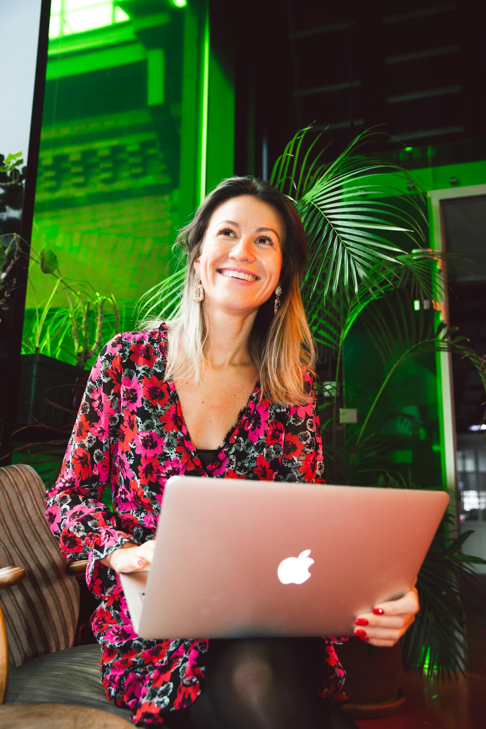 Smiling woman using a laptop in a modern indoor setting with lush greenery.