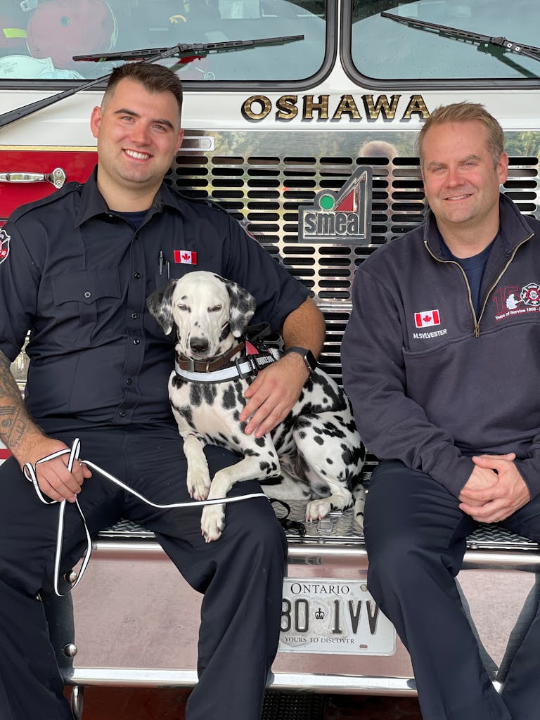 Two firefighters sitting with a Dalmatian dog in front of a fire truck, expressing camaraderie.