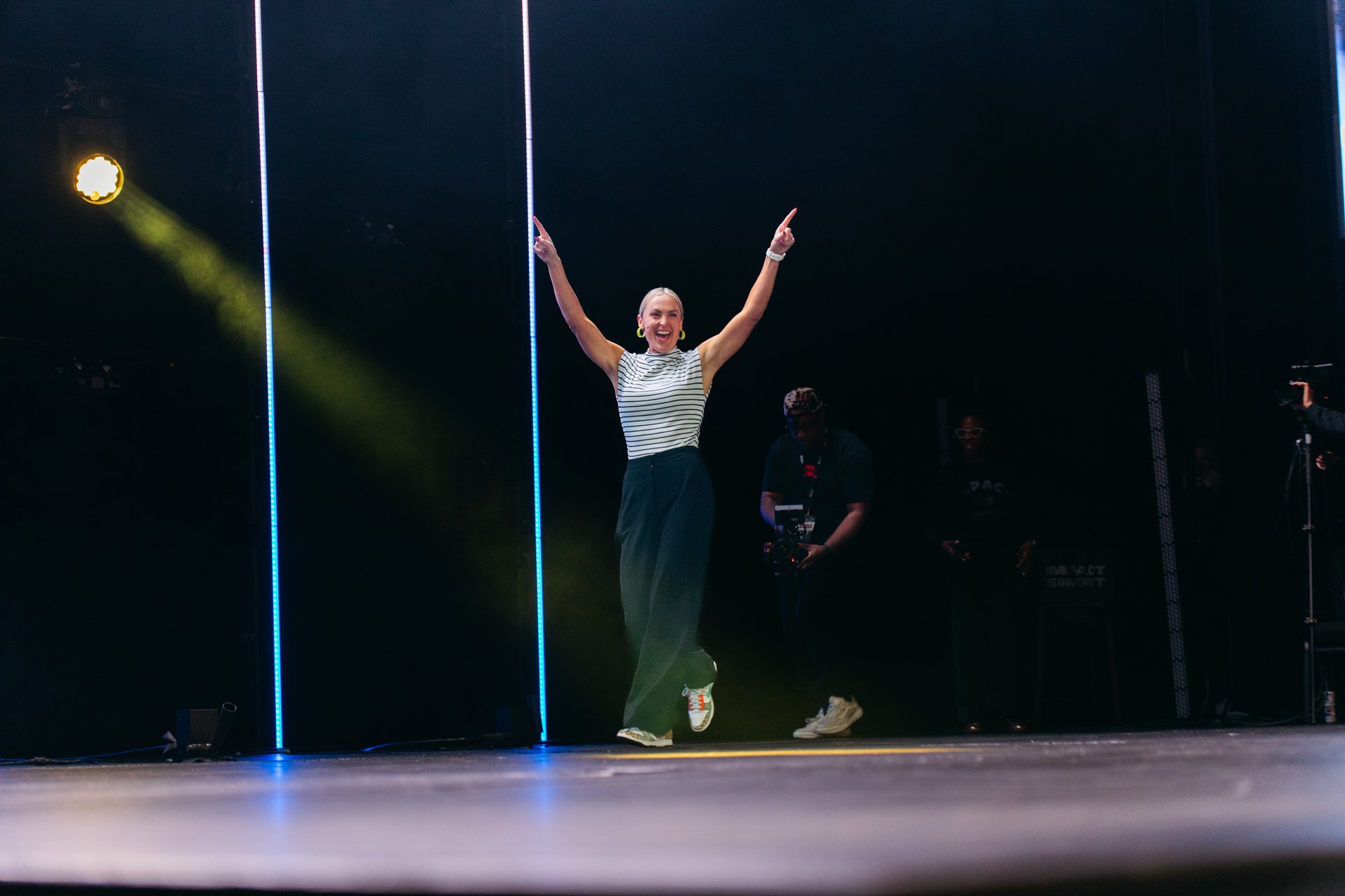 Page Fehling raising both arms on stage under a spotlight, emcee for Catapult's 2027 Leadership and HR Conference